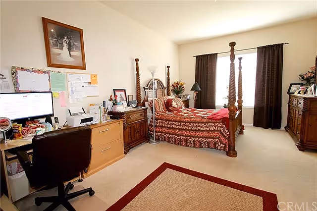 A well-lit bedroom with a four-poster bed covered in a red and brown patterned bedspread. The room includes a wooden dresser, a desk with a computer and office chair, a floor lamp, and a window with dark curtains. Various personal items and framed pictures are placed on the furniture.