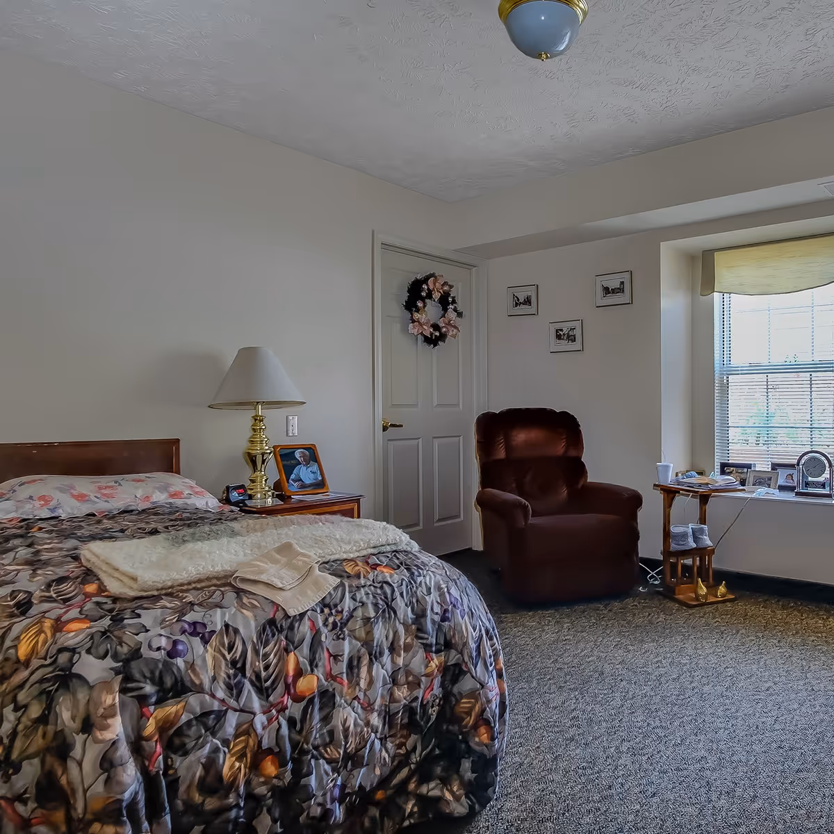 A cozy bedroom in an assisted living facility featuring a bed with a floral patterned comforter, a bedside table with a brass lamp and a framed photo, a comfortable brown armchair, a small wooden side table with various items, and a window with blinds letting in natural light. The walls are decorated with a wreath on the door and three small framed pictures.
