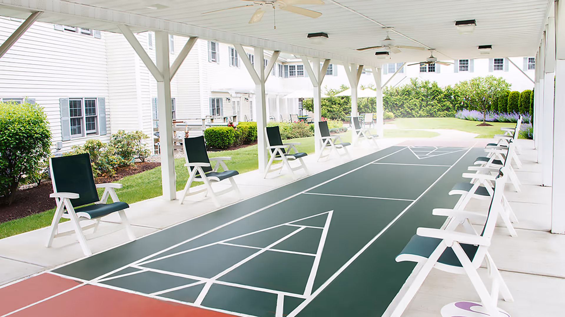 Covered outdoor shuffleboard court with a green playing surface and white chairs lined along both sides under a pavilion next to a senior living building.