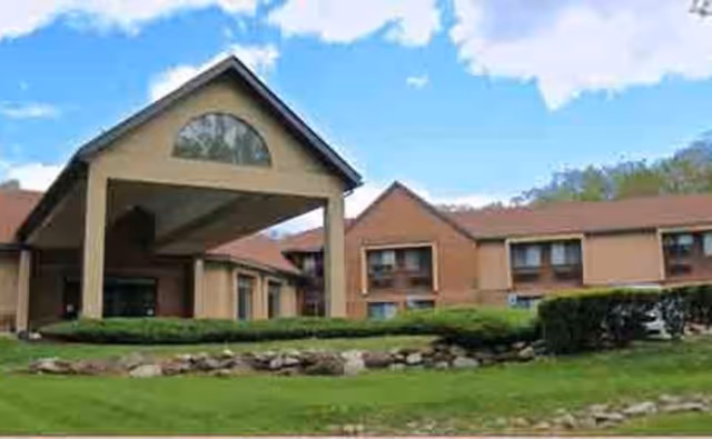 Exterior view of Promenade at Chestnut Ridge senior living facility showing a large covered entrance with a peaked roof, surrounded by well-maintained green lawns, shrubs, and a stone border under a partly cloudy blue sky.