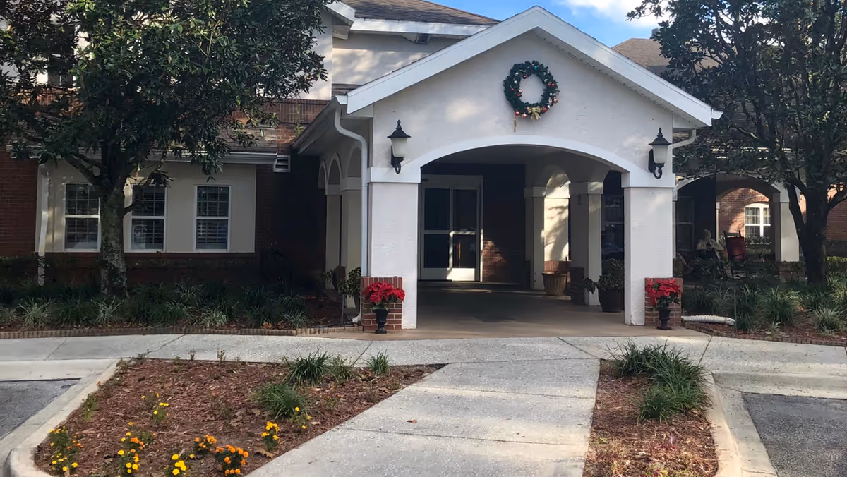 Front entrance of a senior living facility with a covered driveway, decorated with a holiday wreath above the entrance and red poinsettia plants on either side. The building has brick and white exterior walls, with trees and landscaped flower beds in front.