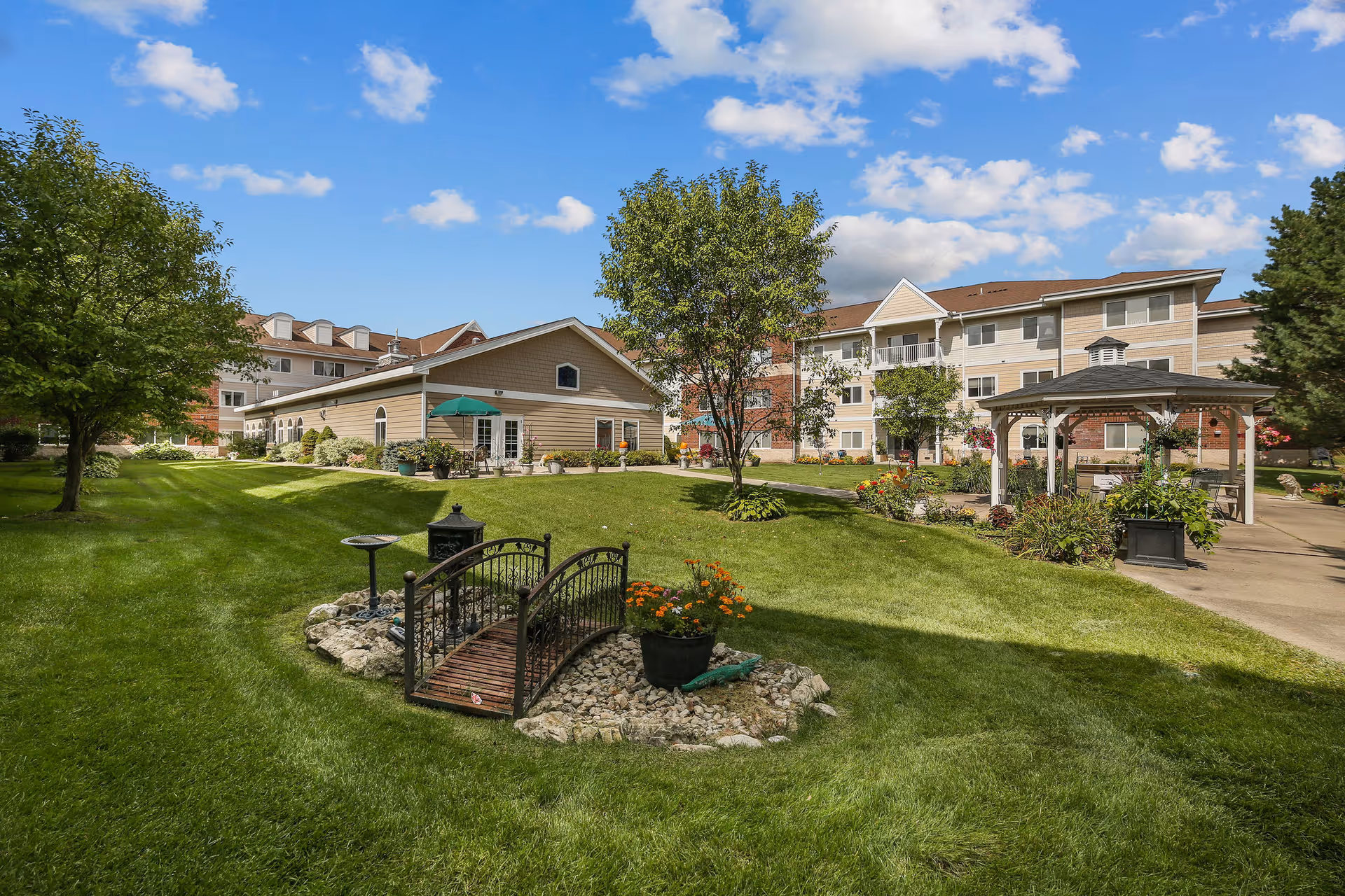 A well-maintained outdoor garden area at The Timbers of Apple Valley featuring a small decorative wooden bridge over a dry rock bed, green grass, trees, colorful flowers, a gazebo with seating, and multi-story residential buildings in the background under a blue sky with scattered clouds.