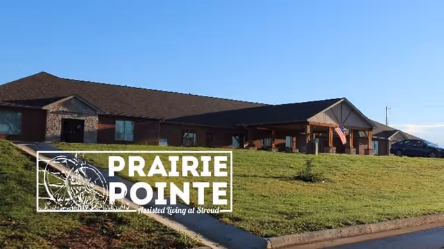 Front exterior of the Prairie Pointe assisted living building with a lawn, covered entrance and an American flag under a clear blue sky.