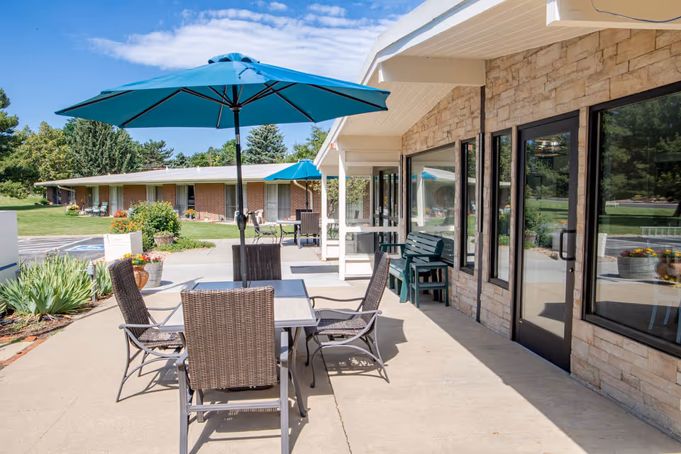 Patio seating with a table, wicker chairs and teal umbrellas on a concrete walkway outside a stone-clad senior living building.