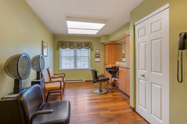 Interior view of a hair salon area in a senior living facility with two black hair dryer chairs on the left, two wooden chairs with orange cushions near a window with blinds and a valance, and a styling station with a black salon chair and mirror on the right. The room has wooden flooring and light green walls.