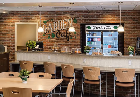Interior view of a cafe area with a brick wall background featuring the sign 'Garden Cafe'. There is a counter with bar stools, a refrigerator labeled 'Fresh Eats' stocked with beverages, and tables with chairs and small potted plants.