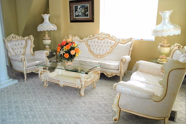 Ornate cream-and-gold upholstered sofa and armchairs arranged around a glass-top coffee table with a floral centerpiece in a bright living room.
