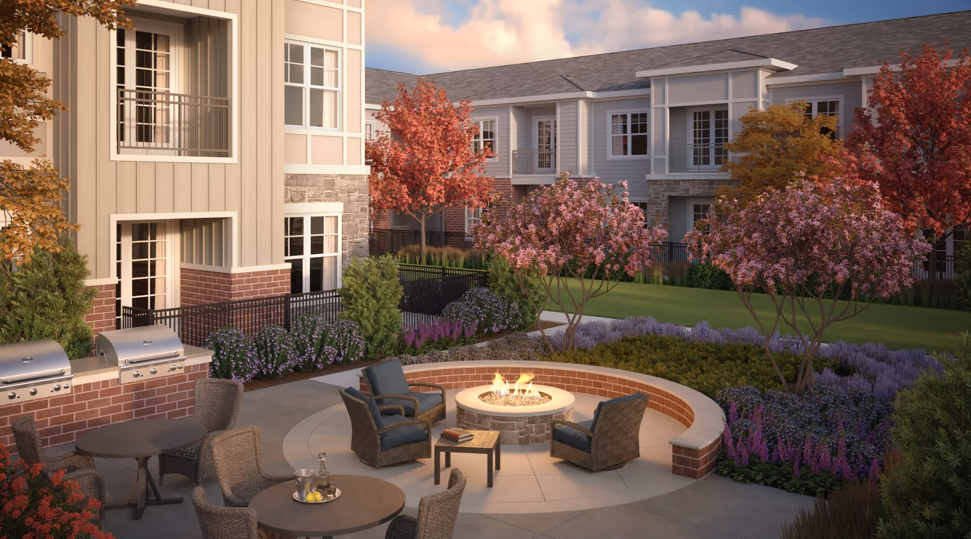 Outdoor courtyard area at Everleigh Naperville featuring a circular fire pit surrounded by cushioned wicker chairs, round tables with chairs, and built-in grills. The space is landscaped with colorful flowering trees and shrubs, with a multi-story residential building in the background under a partly cloudy sky.
