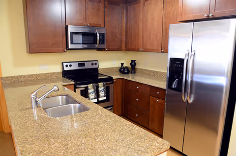 Modern kitchen with granite countertops, stainless steel refrigerator, stove, microwave, double sink, and wooden cabinets.