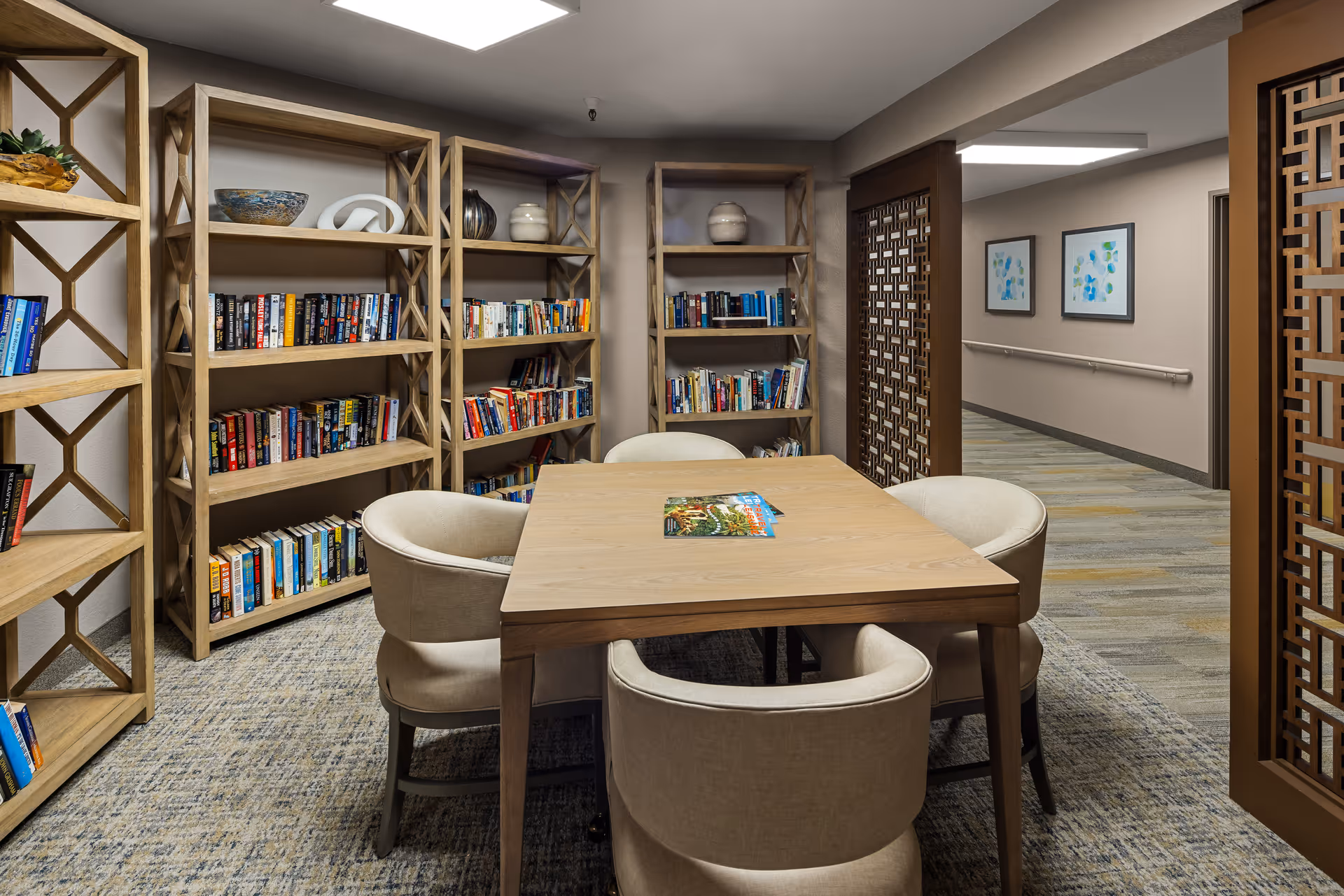 A cozy reading room in a senior living facility featuring wooden bookshelves filled with books and decorative items. In the center, there is a wooden table surrounded by four beige upholstered chairs. The room has a patterned carpet and is softly lit by ceiling lights. Decorative wooden lattice panels partially frame the entrance to the room, and a hallway with framed artwork and handrails is visible in the background.