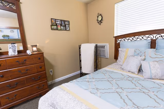 A neatly made bedroom with a wooden headboard and dresser, light blue bedding, and a towel rack by a wall air conditioner.