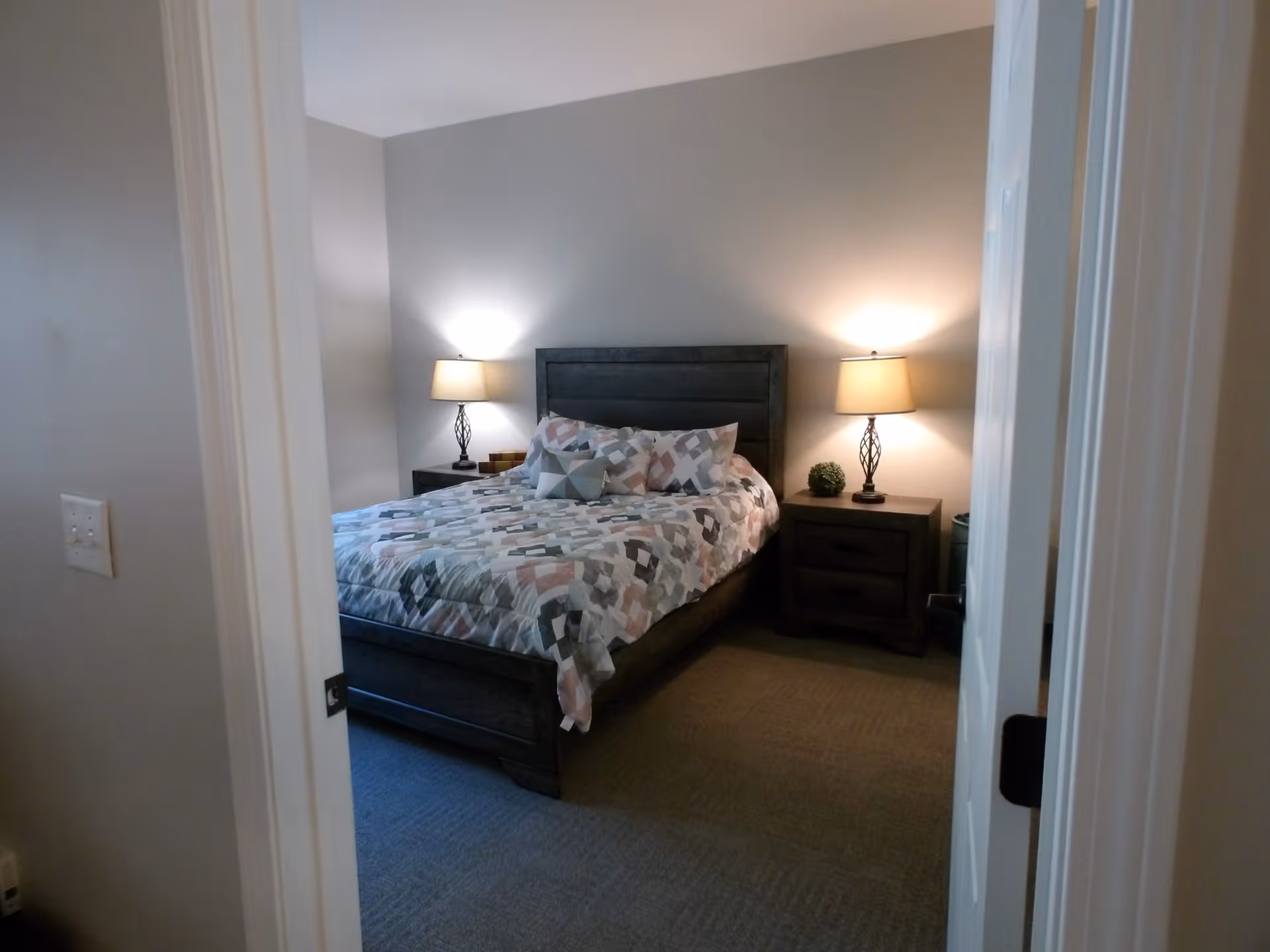 View through a doorway into a bedroom with a bed centered against the far wall. The bed has a patterned quilt and several pillows. On each side of the bed are matching dark wood nightstands with table lamps emitting warm light. The walls are painted light gray and the floor is carpeted.