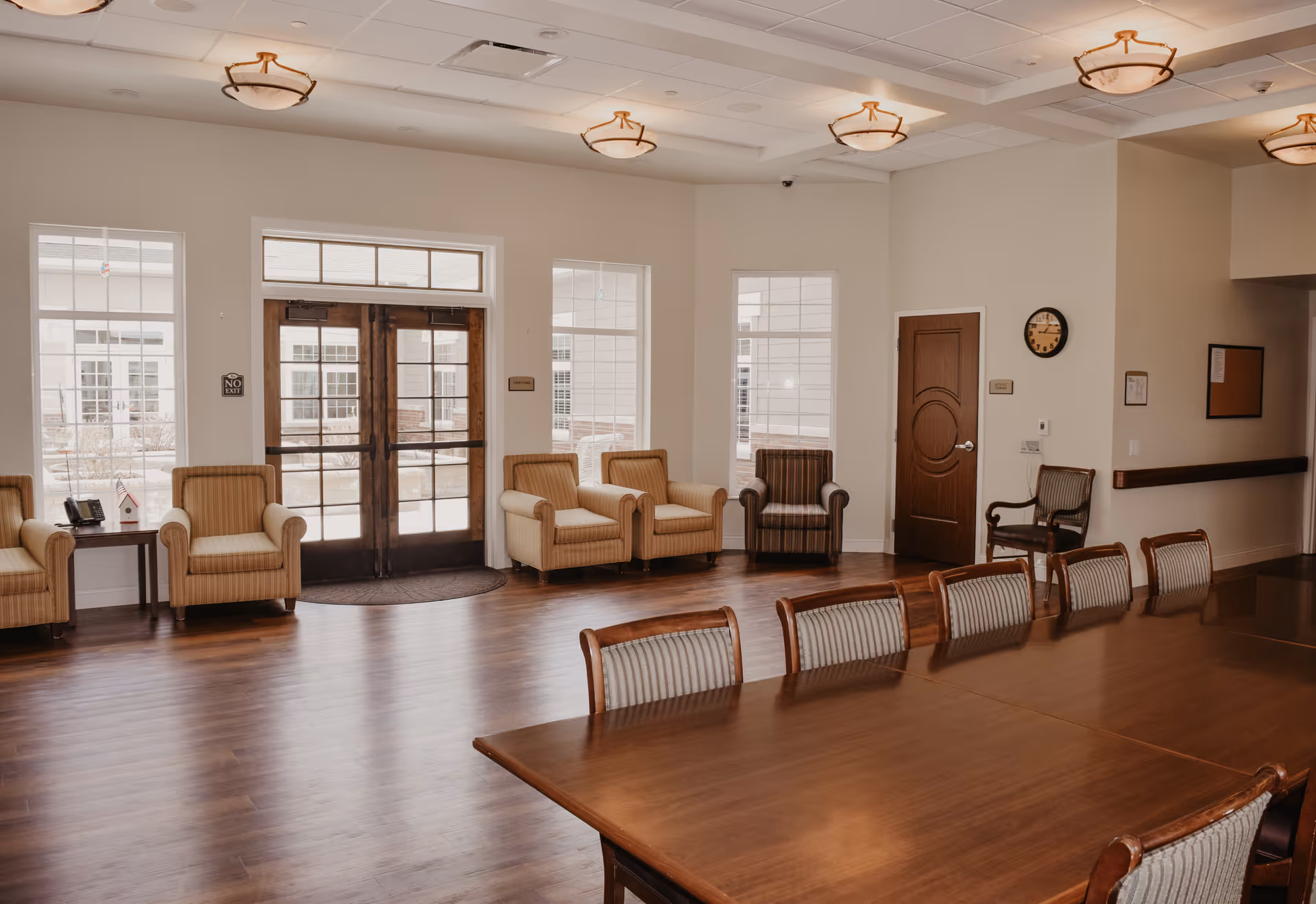 A spacious room with wooden flooring featuring a long wooden table surrounded by chairs with striped upholstery. Along the far wall, there are several armchairs arranged near large windows and a set of double glass doors. The room has a high ceiling with multiple light fixtures and a wall clock above a wooden door.