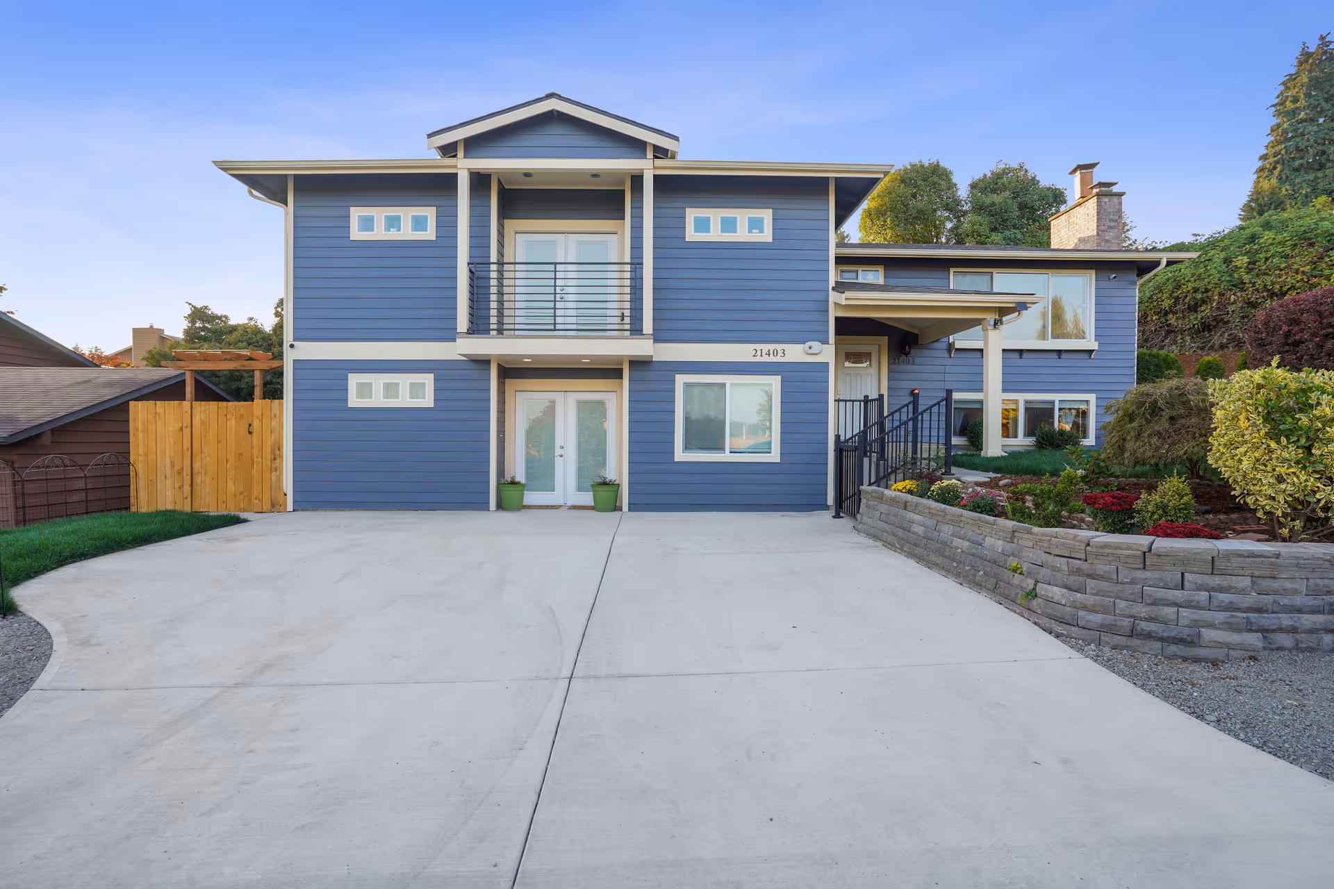 Front exterior view of a two-story blue house with white trim, a concrete driveway, a small balcony above the front door, and landscaped garden beds on the right side.