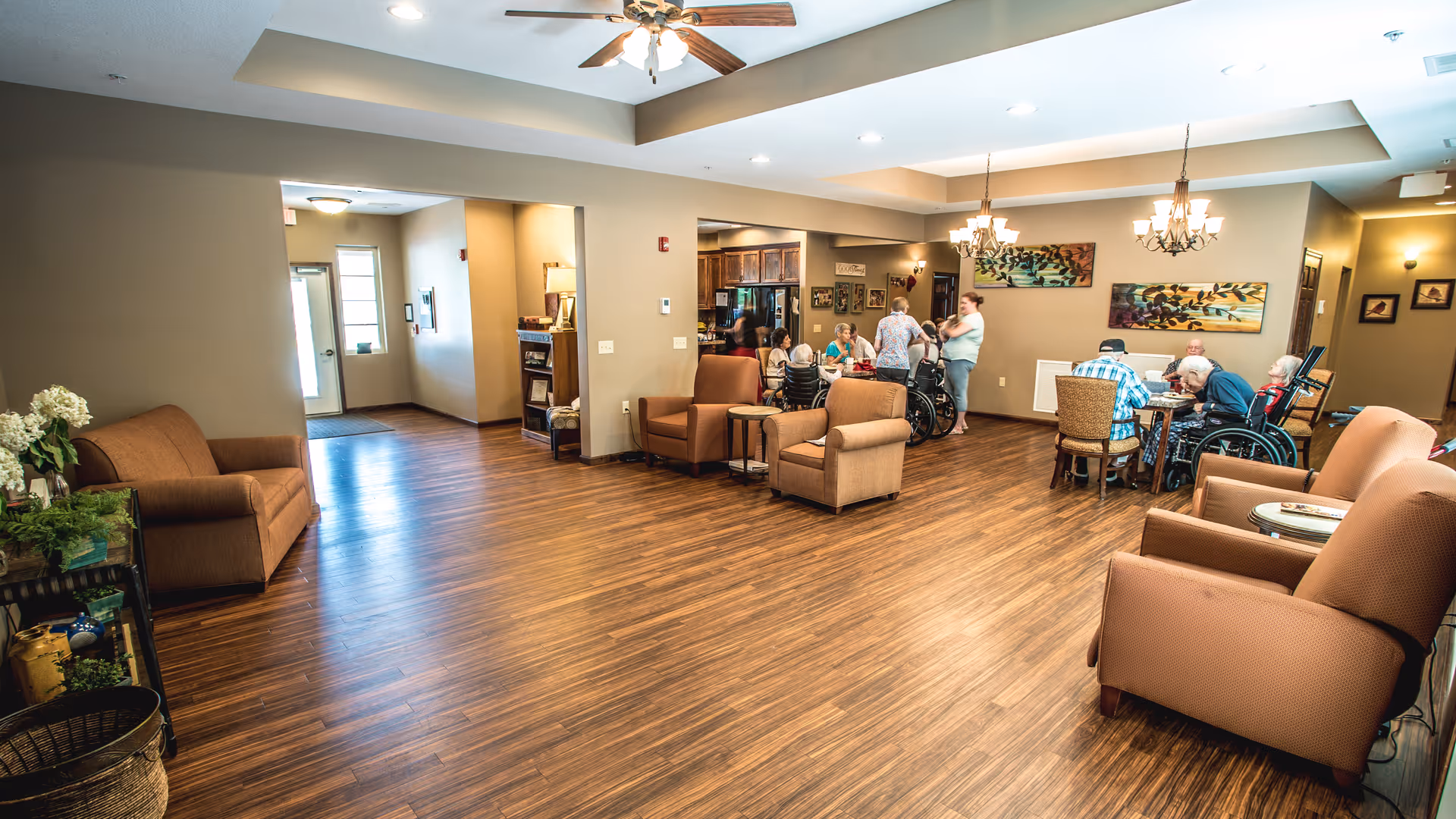 A spacious common area in a senior living facility with wooden flooring and beige walls. Several armchairs and small tables are arranged around the room. In the background, a group of elderly people, some in wheelchairs, are seated around tables, interacting with each other and caregivers. The room is well-lit with ceiling lights and chandeliers, and decorated with framed artwork on the walls.