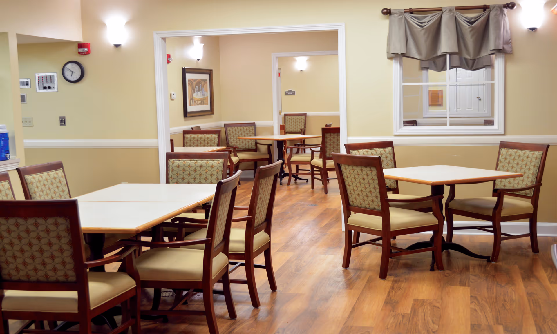 Interior view of a dining area in a senior living facility with multiple wooden tables and cushioned chairs arranged neatly on a wooden floor. The walls are painted beige with white trim, and there are wall-mounted lights and framed artwork. A window with a beige valance is visible on the right side.