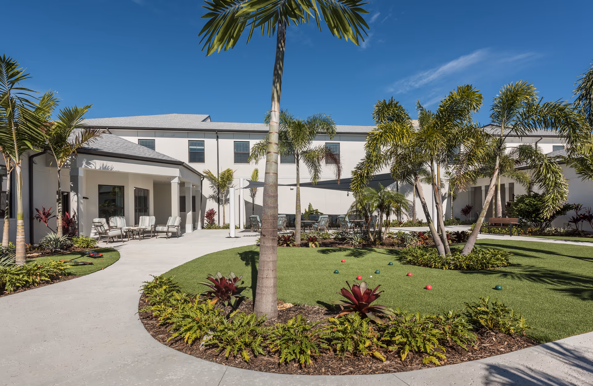 Outdoor courtyard area at HarborChase Of Stuart featuring a well-maintained lawn with bocce balls, surrounded by palm trees and tropical plants. There are several seating areas with chairs and tables under a shaded canopy, and a clear blue sky overhead.