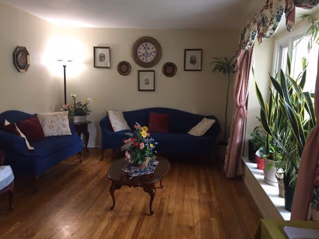 A cozy living room with two blue upholstered sofas adorned with decorative pillows, a wooden coffee table with a floral centerpiece, hardwood flooring, a floor lamp, framed wall art, and a window with floral curtains and several potted plants on the windowsill.