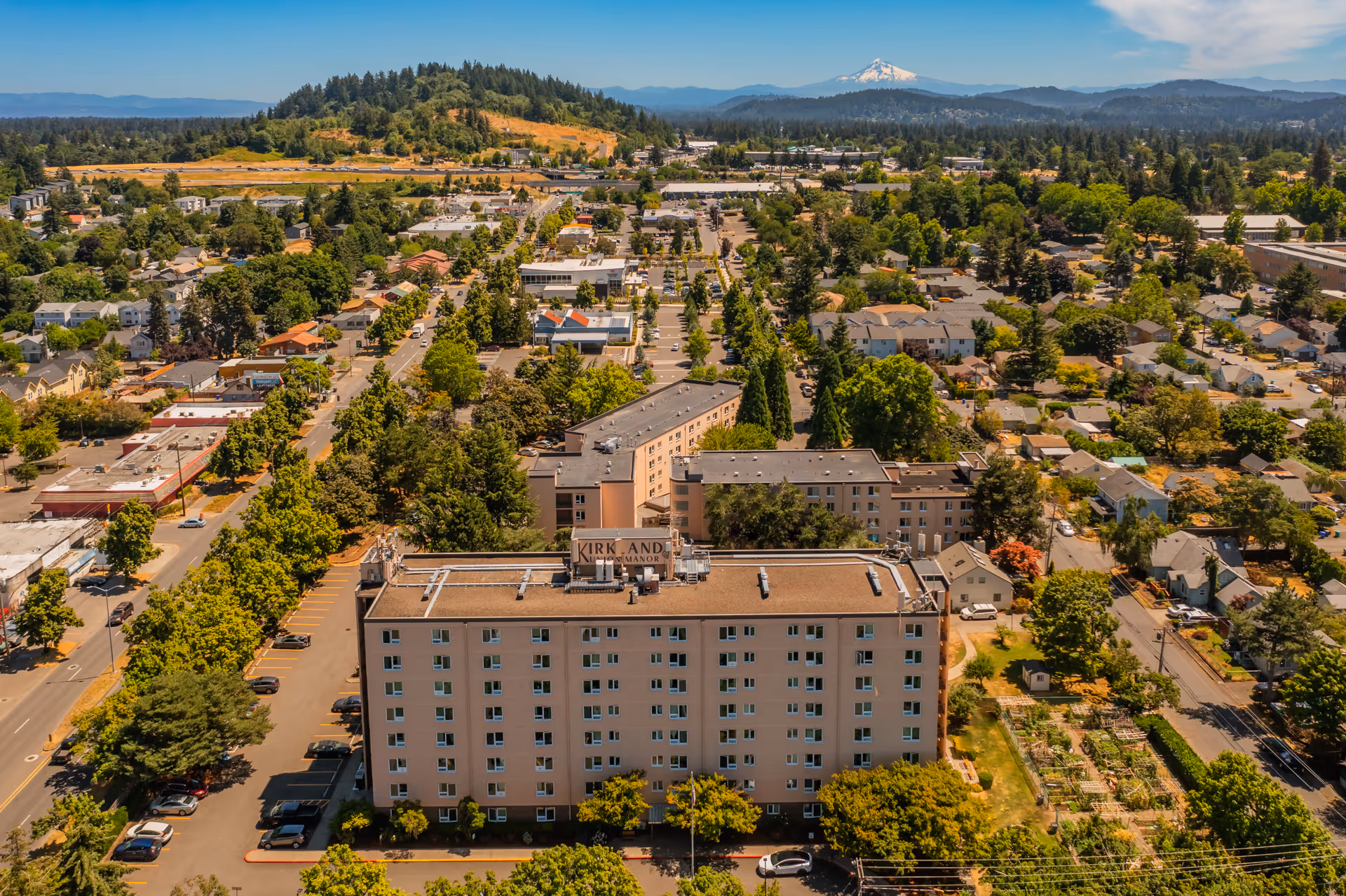Aerial view of Kirkland Union Manors, a multi-story residential building surrounded by trees and other smaller buildings in a suburban area with a mountain visible in the background under a clear blue sky.