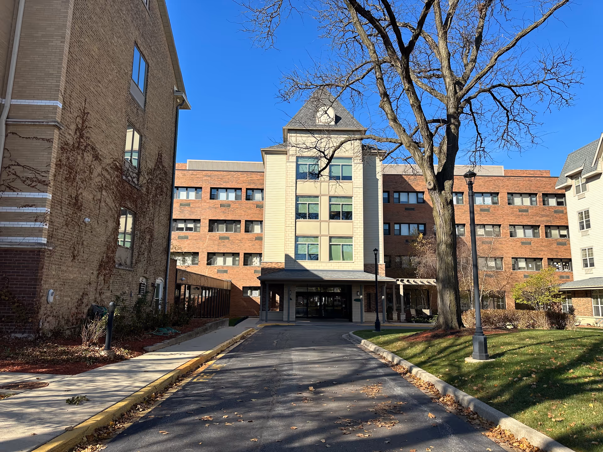 Exterior view of a senior living facility named Norwood Crossing, showing a multi-story brick and beige building with a peaked roof section in the center. There is a driveway leading to the entrance, a large leafless tree on the right, and a clear blue sky above.