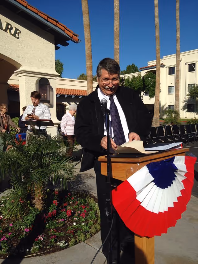 A man in a black jacket and tie stands at a wooden podium decorated with a red, white, and blue fan-shaped banner, speaking into a microphone outdoors. Behind him are several people, palm trees, and a building with a tiled roof under a clear blue sky.