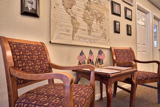 Two wooden chairs with patterned cushions are placed on either side of a small wooden table holding three small American flags and some papers. Behind the chairs, a wall displays a large world map and several framed photographs. A white door is visible in the background.
