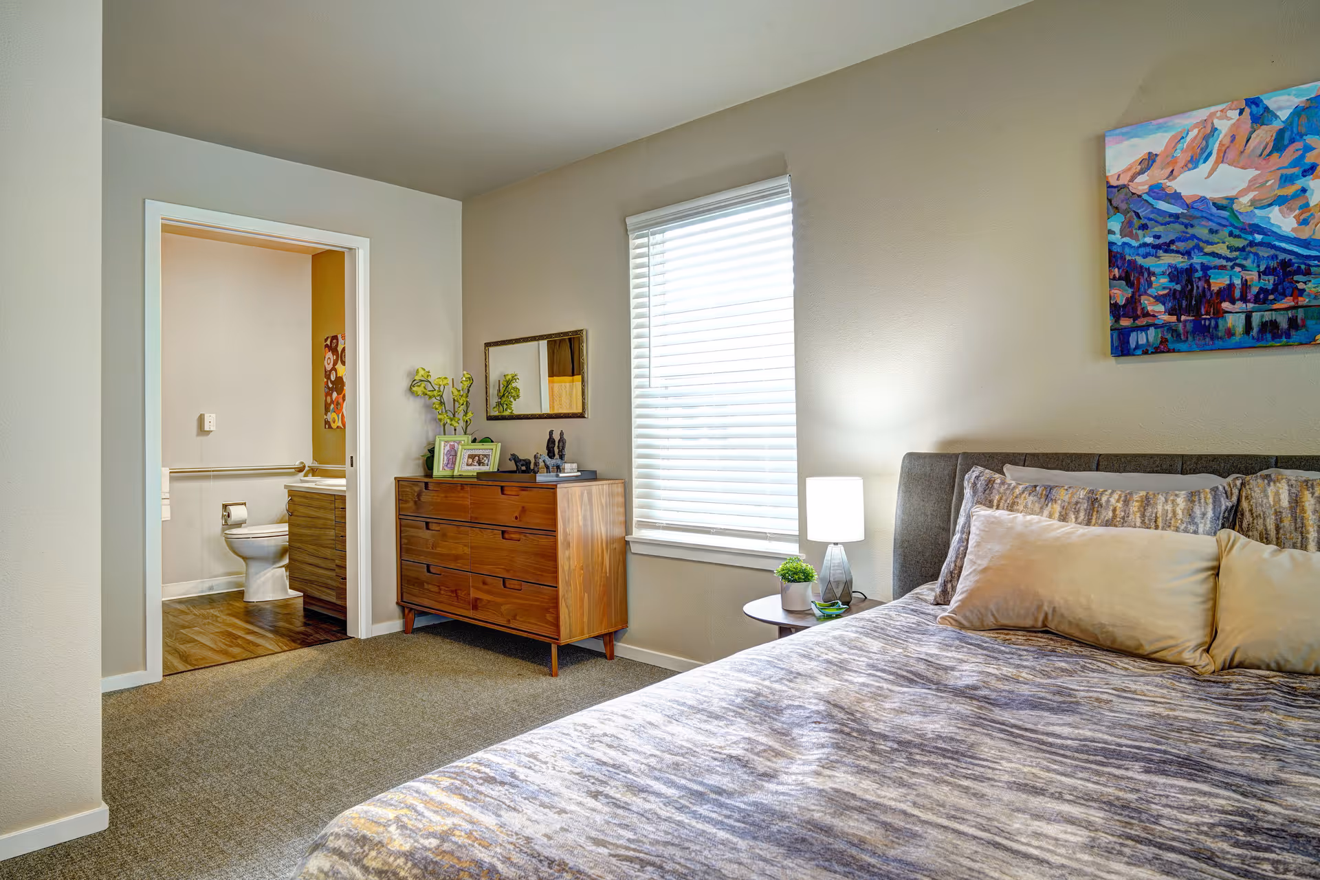 A bedroom with a bed covered in a patterned comforter and multiple pillows. Next to the bed is a small round side table with a lamp and a small plant. A window with white blinds is on the wall behind the table. Across from the bed is a wooden dresser with framed photos, decorative items, and a mirror above it. An open doorway leads to a bathroom where a toilet and part of a vanity are visible.