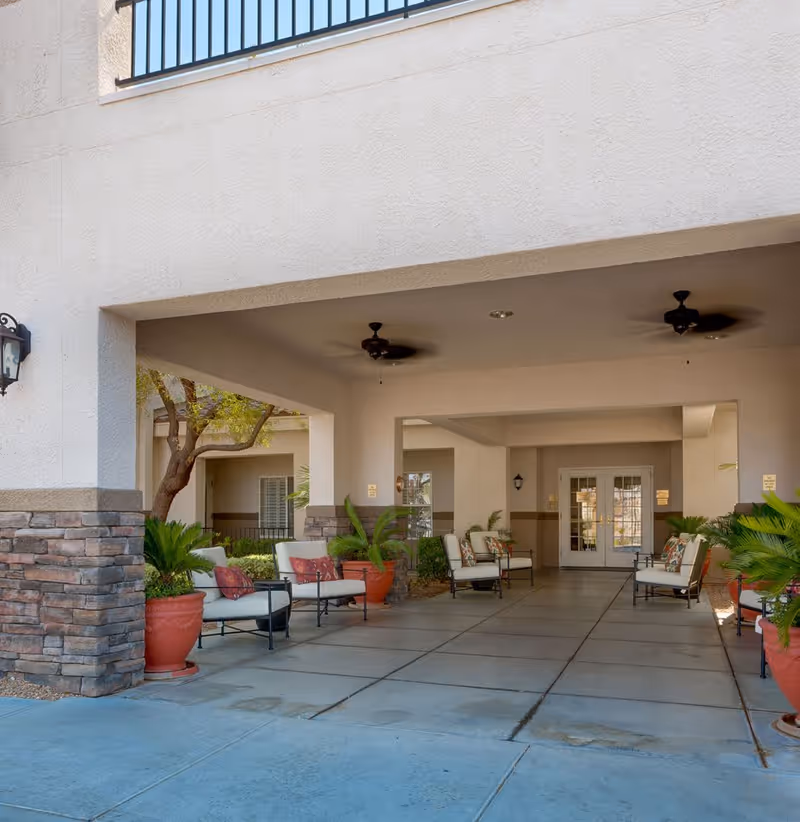 Covered outdoor seating area with cushioned chairs and potted plants, ceiling fans, and a double door entrance in the background at a senior living facility.