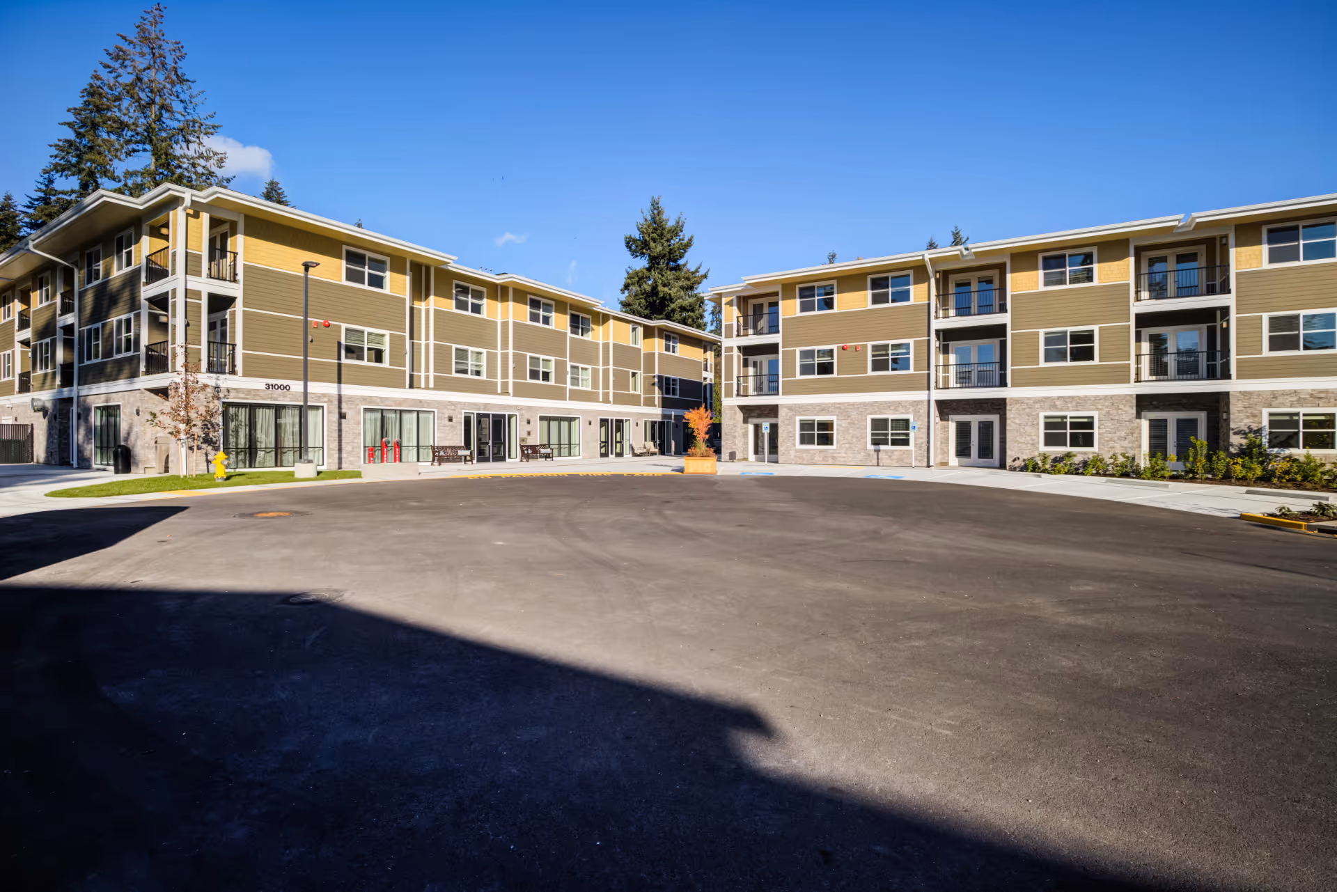 Exterior view of a senior living community building with three stories, featuring large windows, balconies, and a paved driveway area in front. The building has a combination of yellow and olive green siding with stone accents on the lower level. There are benches and a fire hydrant near the entrance, with trees and a clear blue sky in the background.