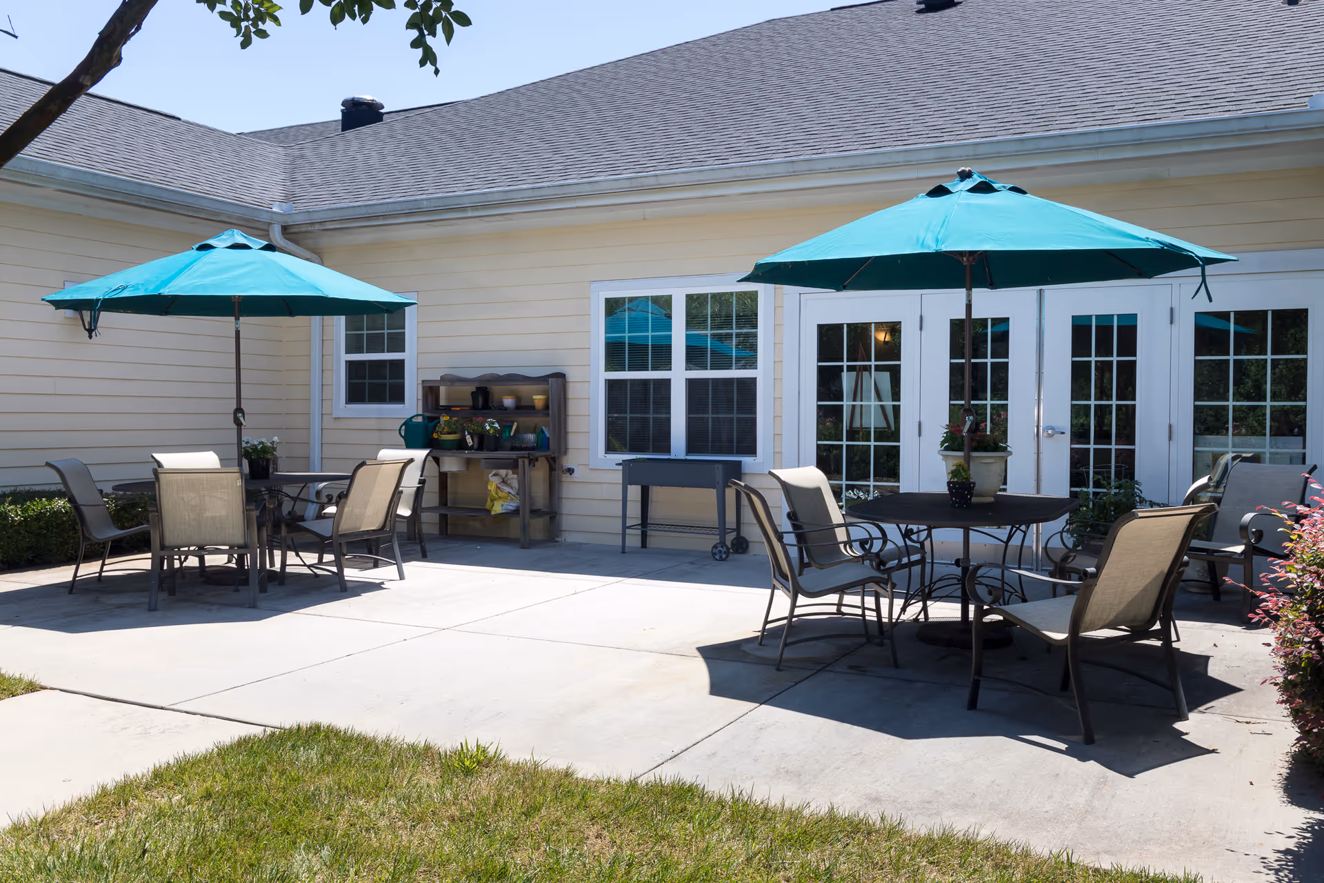 Outdoor patio area at Cadence at Clemmons featuring two round tables with chairs and teal umbrellas providing shade. The patio is adjacent to a light yellow building with white-framed windows and French doors. There is a small shelving unit with gardening supplies and a grill on the patio.