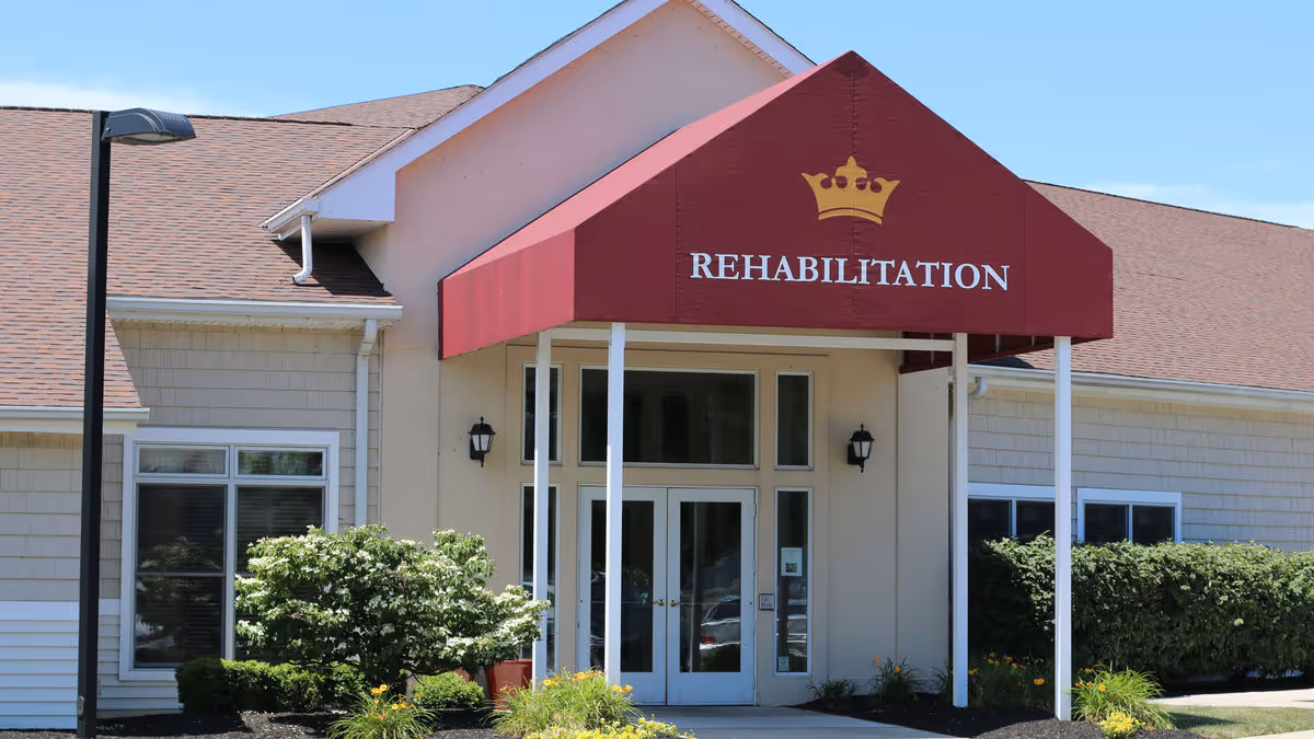 Front entrance of a senior living facility with a red canopy reading "REHABILITATION" over double glass doors and surrounding shrubs.