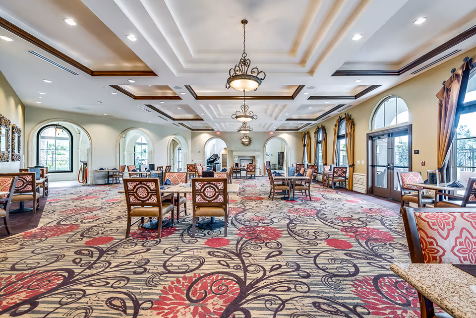 Spacious dining room in a senior living facility with patterned carpet, multiple tables and chairs, large windows with curtains, and elegant ceiling lights. The room features arched doorways and a fireplace at the far end.
