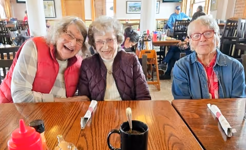 Three elderly women sitting at a wooden dining table in a bright dining room, smiling at the camera. The table has a black coffee mug, a red condiment bottle, and wrapped utensils. Other people are visible in the background seated at tables.