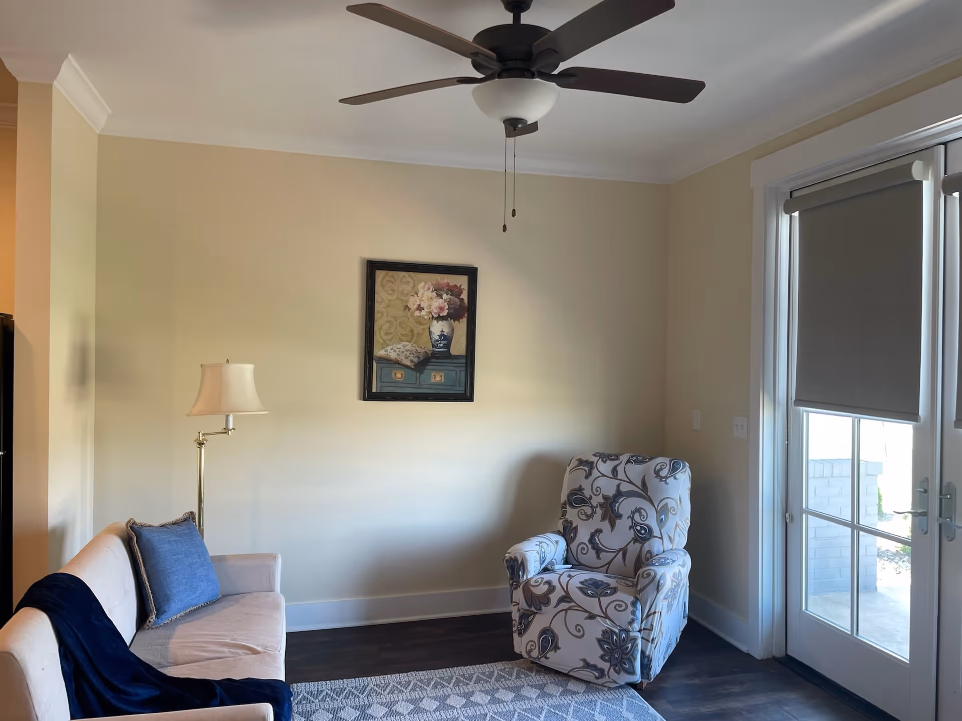 A cozy living room with a beige sofa adorned with a blue pillow and a dark blue throw blanket, a floor lamp with a white shade, a patterned armchair, a ceiling fan with a light fixture, a framed floral painting on the wall, and glass doors with gray roller blinds leading outside.