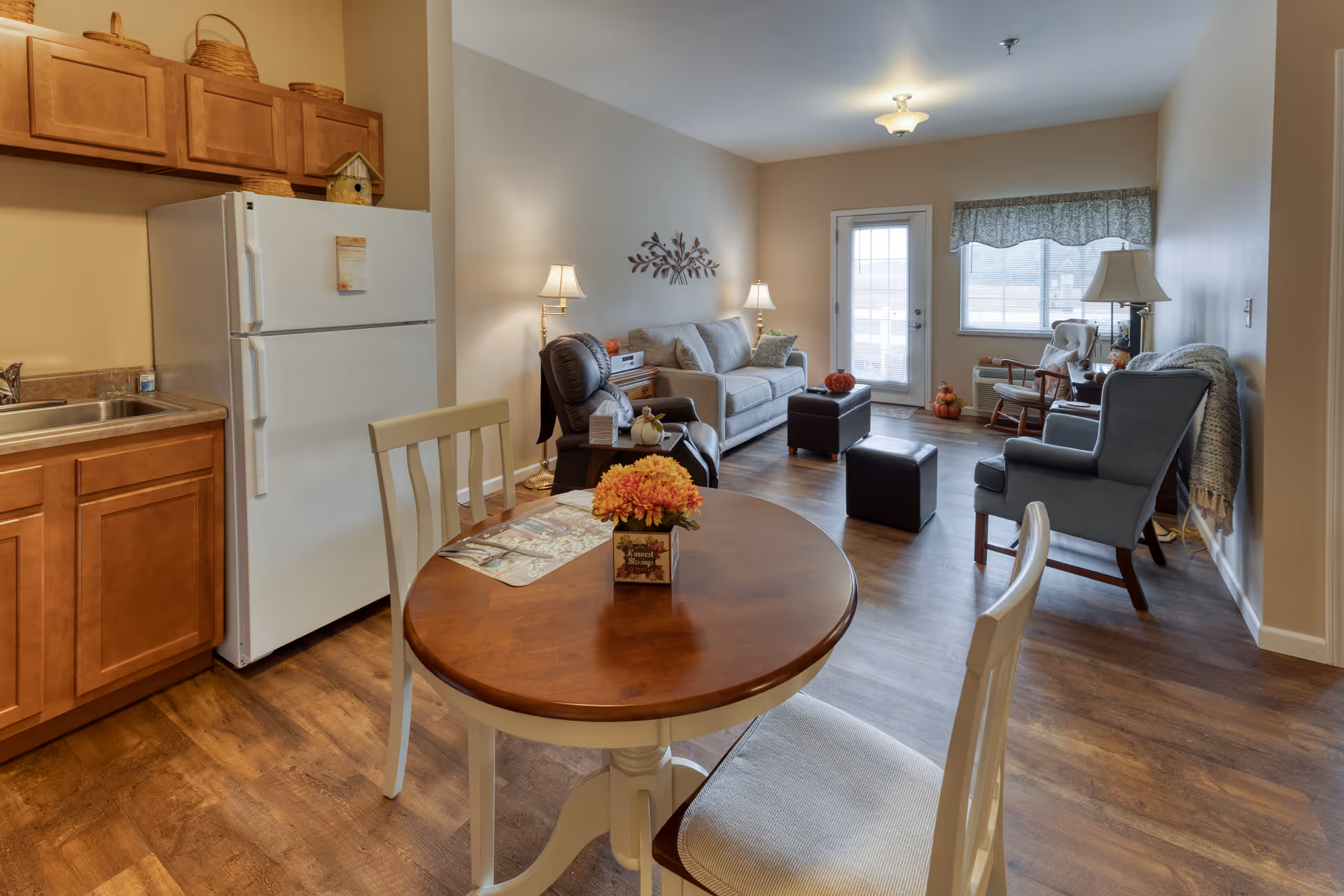 A cozy senior living apartment interior featuring a small kitchen area with wooden cabinets, a white refrigerator, and a sink. In the foreground, there is a round wooden dining table with two chairs and a flower centerpiece. The living area includes a beige sofa, a recliner chair, two armchairs, a small ottoman, and several lamps providing warm lighting. A door with glass panels and a window with a valance allow natural light into the room. The floor is wood-style laminate.