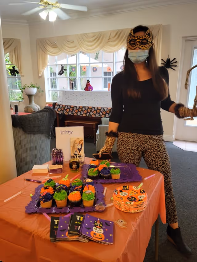 A woman dressed in a leopard print costume with a matching mask and gloves stands next to a table decorated for Halloween. The table is covered with an orange tablecloth and has trays of Halloween-themed cupcakes, festive napkins, plates, and some promotional materials. The room has large windows with cream-colored curtains and Halloween decorations on the windows and walls.