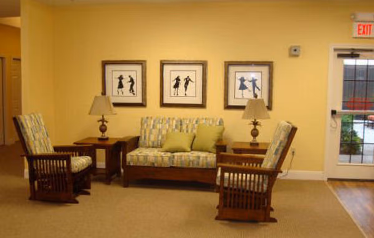 A cozy seating area in a living room with a wooden loveseat and two matching armchairs upholstered in a patterned fabric. There are two wooden side tables with lamps on either side of the loveseat. Three framed silhouette art pieces hang on the yellow wall behind the seating. A glass door with an exit sign above it is visible to the right.