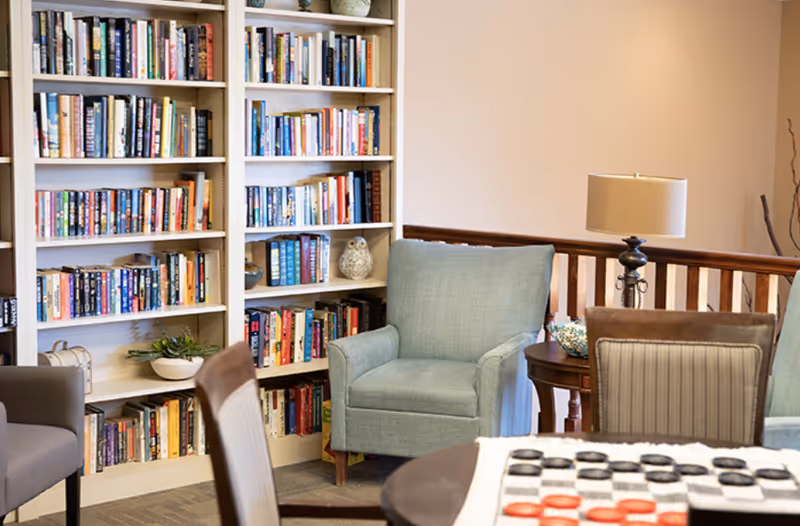 A cozy reading and sitting area with a light blue armchair, a wooden side table with a lamp, and bookshelves filled with books. In the foreground, there is a table with a checkers game set up. The room has a warm and inviting atmosphere.