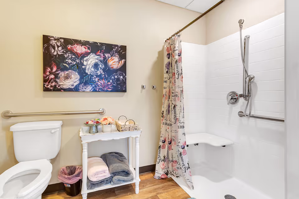 A senior living facility bathroom featuring a white toilet with a grab bar on the wall beside it, a small white table holding folded towels and decorative items, and a walk-in shower with a floral-patterned curtain, a wall-mounted shower seat, and grab bars. A floral painting hangs on the wall above the toilet.