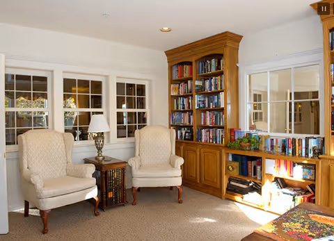 A cozy sitting area with two beige upholstered armchairs placed around a small wooden side table with a lamp. Behind the chairs are windows with white frames. To the right, there is a large wooden built-in bookshelf filled with books and some decorative items. Sunlight streams in from the right side, illuminating part of the bookshelf and carpeted floor.
