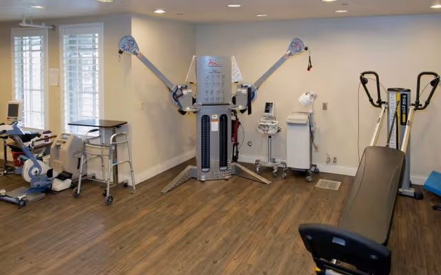 A room with various physical therapy and exercise equipment including a multi-functional exercise machine, a stationary bike, a walker, a blood pressure monitor, and other medical devices, with wood flooring and white walls.