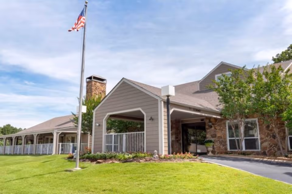 Exterior view of Vitality Living Pleasant Hills facility showing a covered entrance with stone and siding walls, a driveway, well-maintained green lawn, trees, and an American flag on a flagpole against a partly cloudy sky.