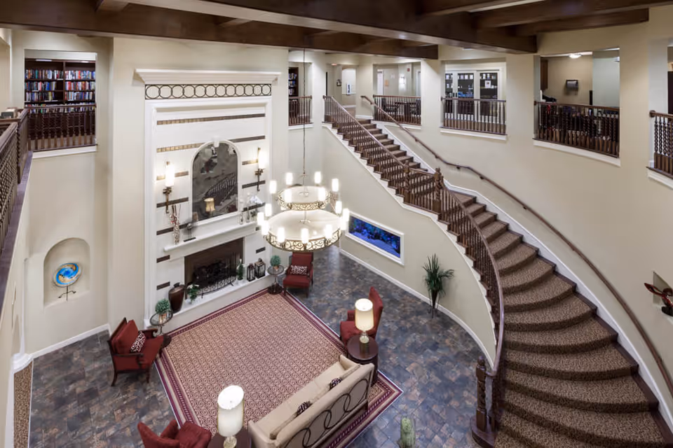 Spacious two-story living room area with a large fireplace, a patterned rug, multiple chairs and a sofa, a curved staircase with wooden railings, a large chandelier hanging from the ceiling, and bookshelves on the upper level.