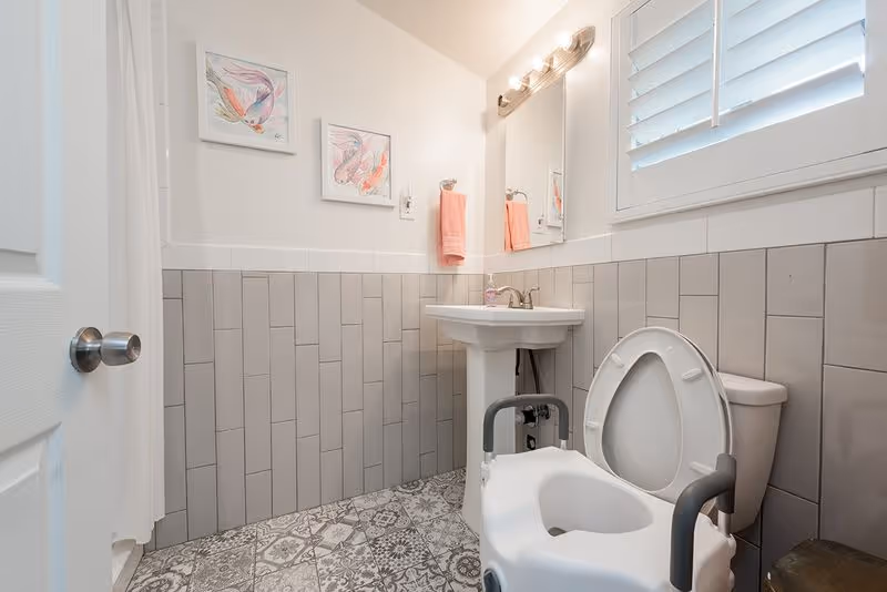 A clean bathroom with a white toilet equipped with safety armrests, a white pedestal sink with a soap dispenser, a large mirror with lights above it, and two framed fish paintings on the wall. The walls are partially tiled with vertical gray tiles, and the floor has patterned tiles. A pink hand towel hangs on a ring near the sink, and a window with white shutters is visible.