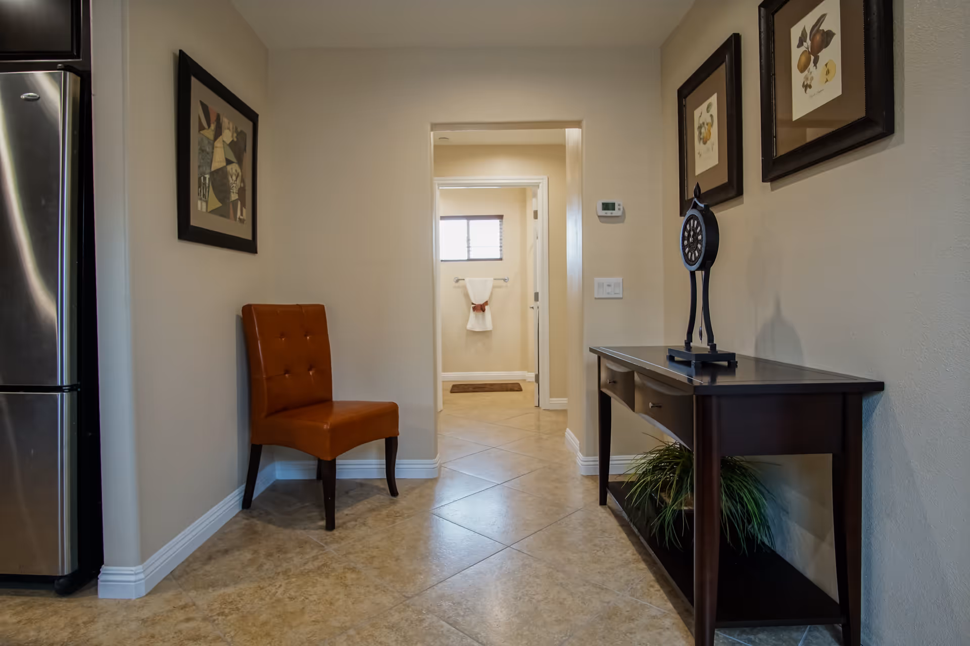 Interior hallway with beige tiled floor and cream-colored walls. On the left, there is a brown leather chair and a framed abstract painting above it. On the right, a dark wooden console table with two drawers holds a decorative clock and a plant underneath. At the end of the hallway, a doorway leads to a bathroom with a towel hanging on a rack beneath a small window.