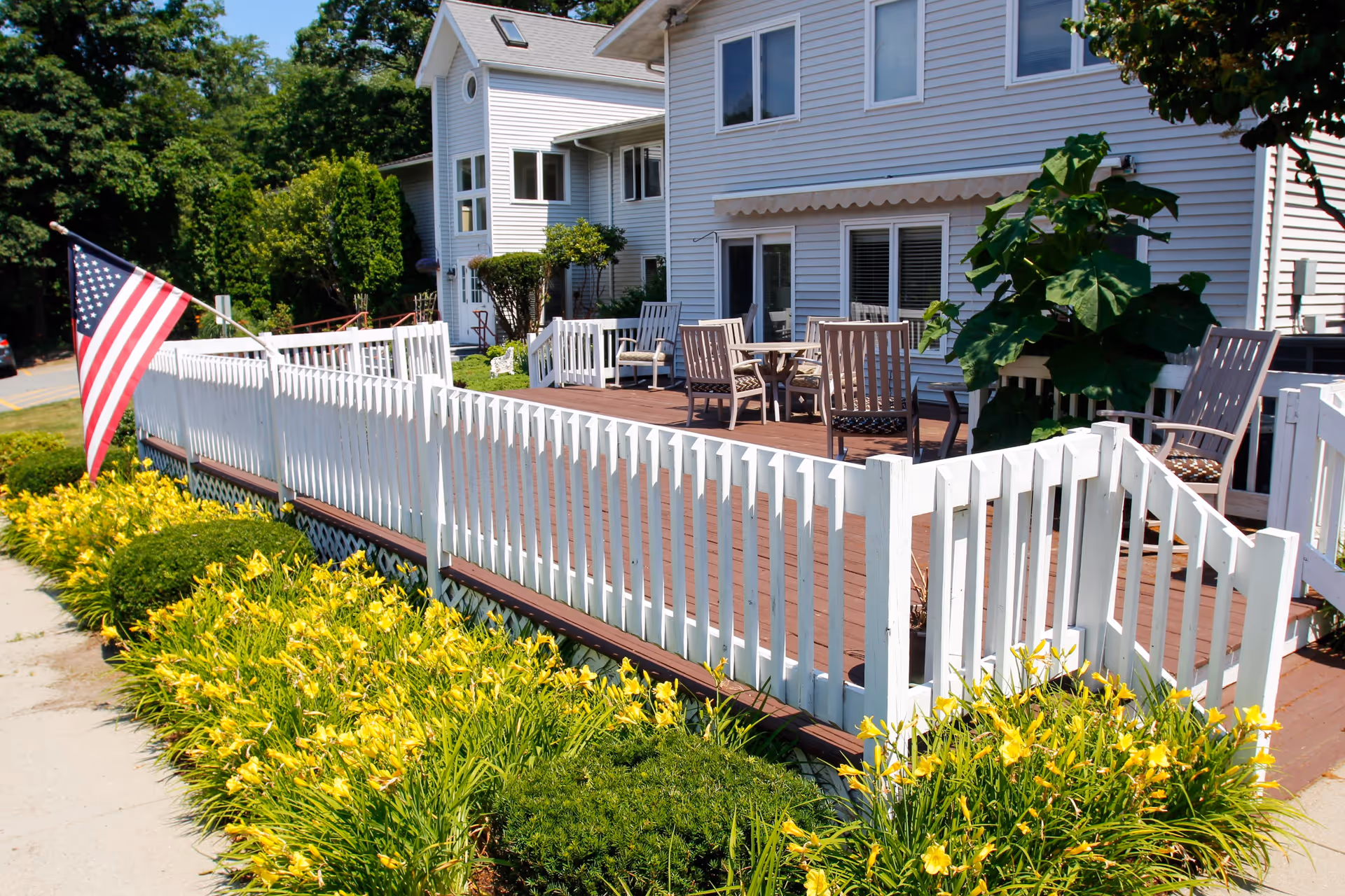 Outdoor patio area of a senior living facility with white wooden railing, several chairs and tables on a wooden deck, an American flag, and yellow flowering plants along the sidewalk.