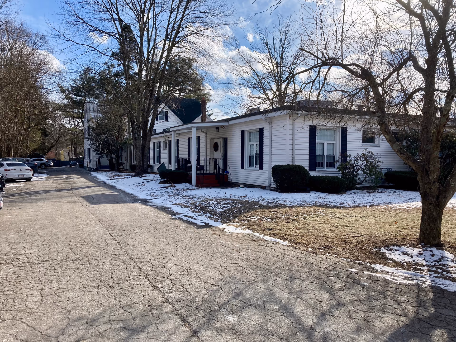 Single-story white building with a covered porch, parking area, leafless trees, and patches of snow on the ground.