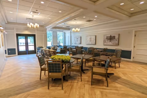 A spacious dining room with multiple wooden tables and chairs arranged neatly. The room features large windows allowing natural light to enter, light-colored wooden flooring, and decorative wall art. Ceiling lights and chandeliers provide additional lighting, and there is a double door leading outside.