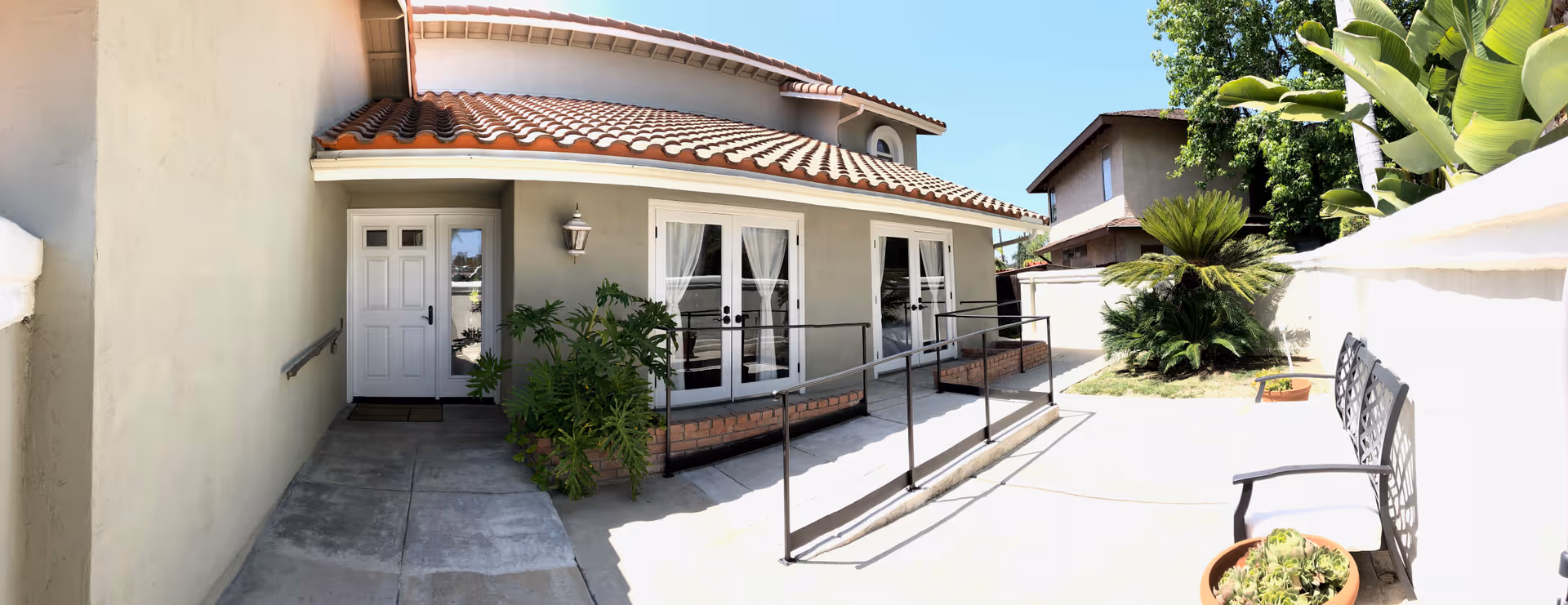 Outdoor view of a single-story building with a tiled roof and light-colored walls. There is a white door and two large windows with white curtains. A concrete ramp with black metal railings leads to the entrance. On the right side, there is a small garden area with green plants and a bench with a potted plant nearby. The sky is clear and blue.