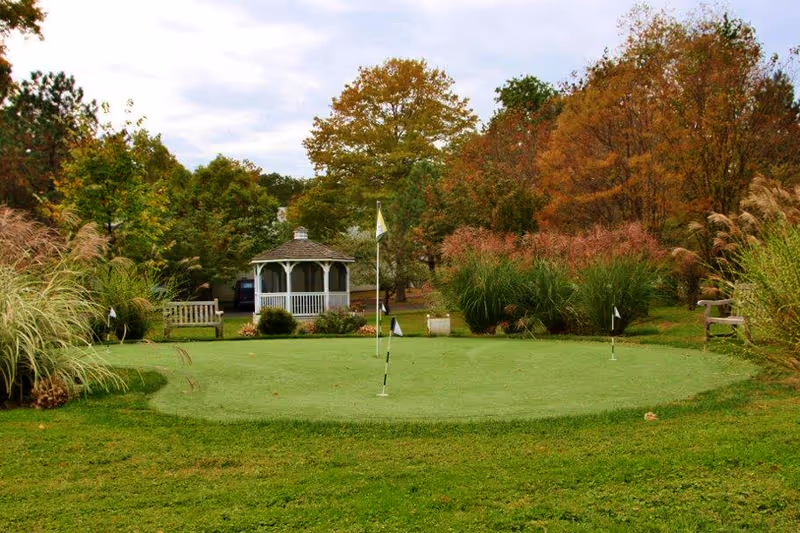 A small putting green with flags surrounded by benches, ornamental grasses, autumn trees, and a white gazebo in the background.